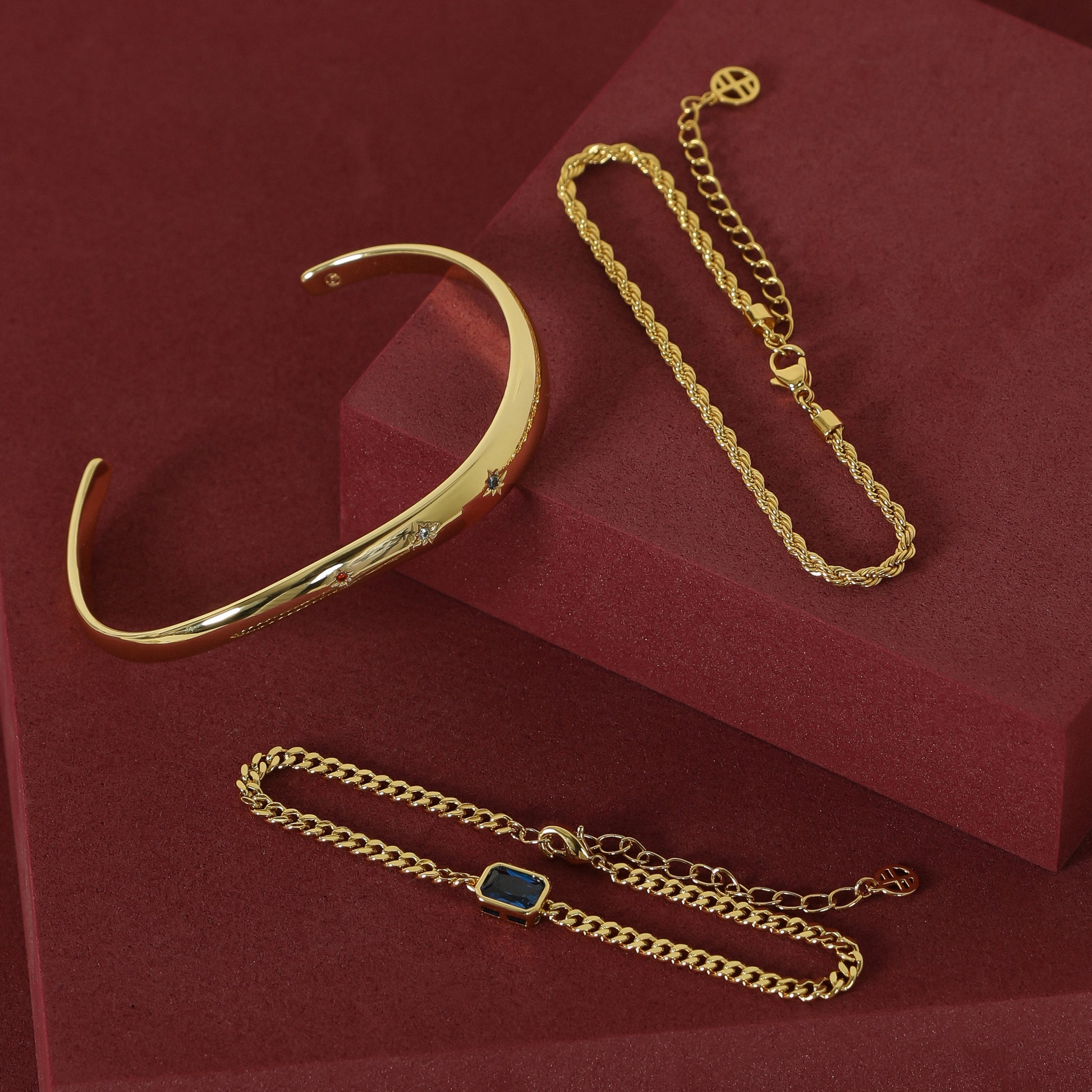 gold chain bracelet with a navy blue cubic zirconia asymmetrical cuff and rope bracelet set on red background 