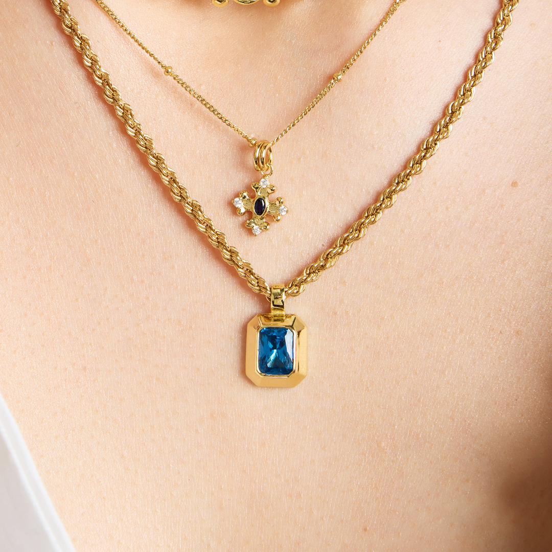 Gold charm with Blue emerald cut cubic zirconia bezel set Inspired by the halls of Versailles on rope chain, layered with bobble chain and The Versailles Charm features the intricate fleur-de-lis details on neck