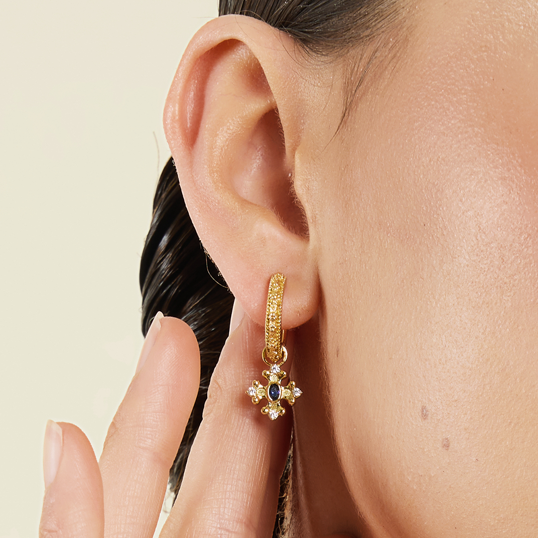 gold charm that features the intricate fleur-de-lis details found in the iconic Palace of Versailles accompanied by the deep blue stone on gold hoops close up in ear 
